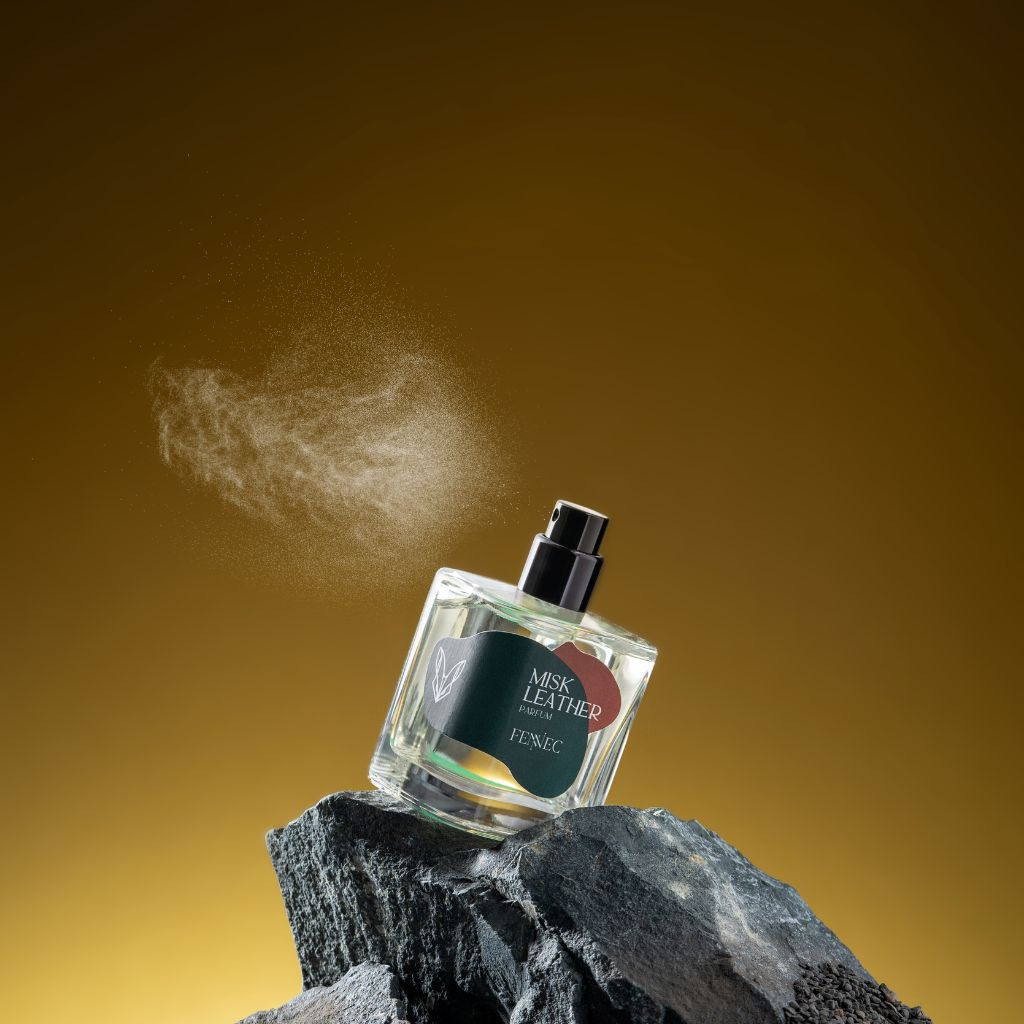 A bottle of Fennec's Misk Leather Parfum rests on a jagged rock with swirling mist, set against a smooth amber gradient that recalls the warm tones of the North African desert fox’s habitat.
