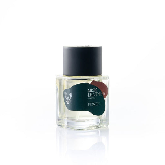 A clear glass bottle of Misk Leather Parfum by Fennec features a black cap and a white label with abstract green and red shapes plus a small outline of the North African fennec fox, the brand's namesake.