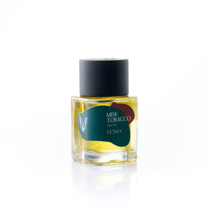 A clear glass perfume bottle with yellow liquid, labeled Misk Tobacco Parfum by Fennec, has a black cap and a green and red abstract label inspired by North African desert colors, displayed against a white background.