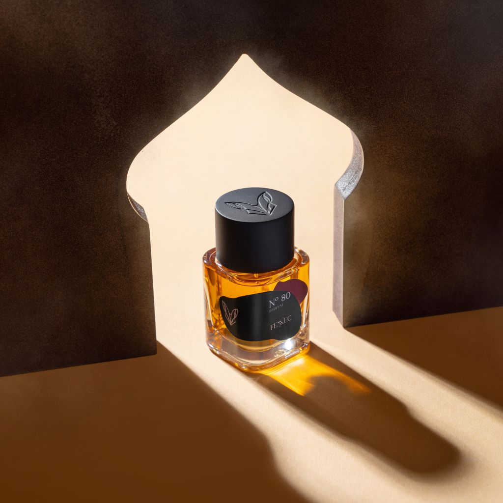 A bottle of Fennec N80 Parfum, with amber liquid, rests in a beam of light within an arched cutout casting dramatic shadows on beige. The black-capped bottle has a black label with gold details, evoking the allure of North Africa’s desert fox.