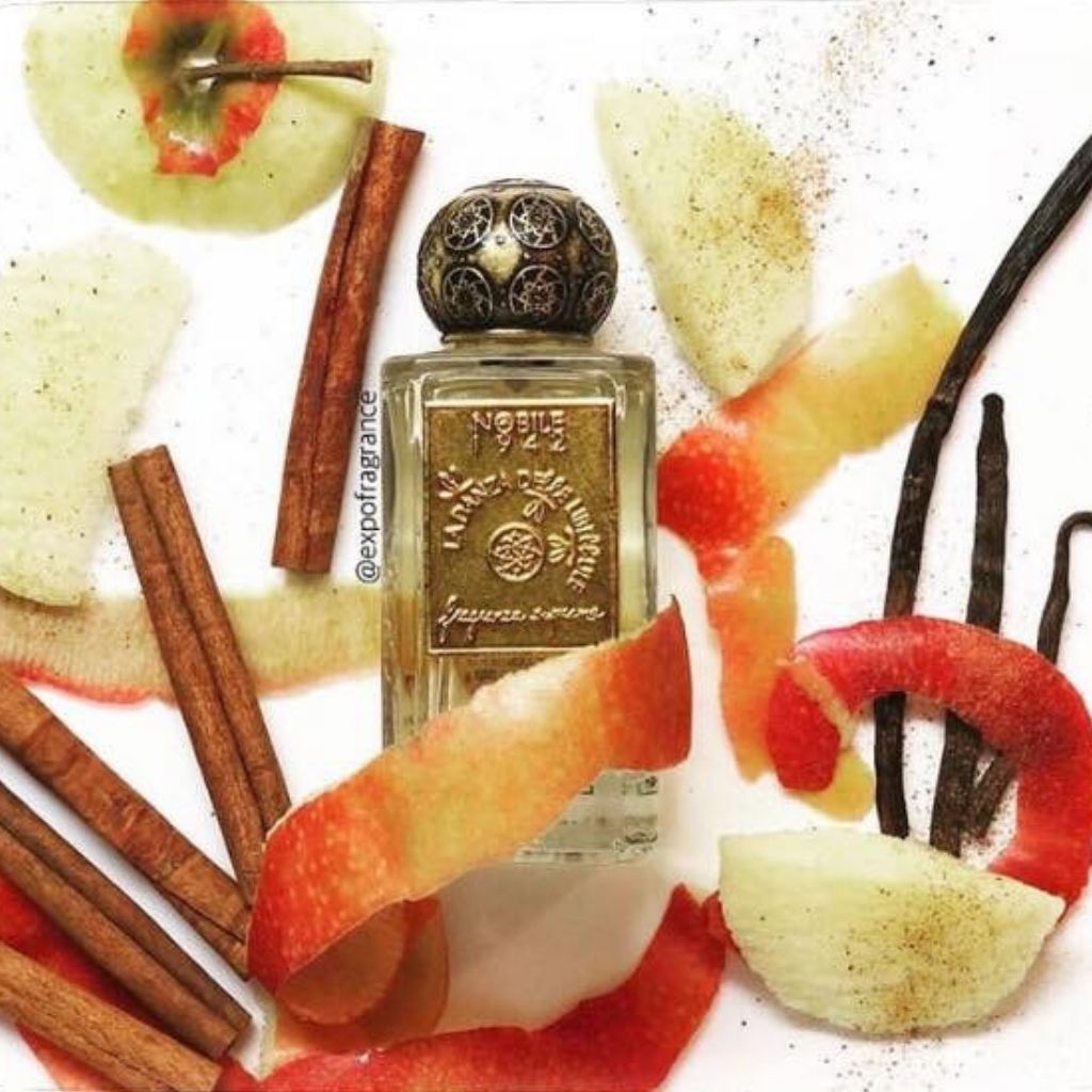 A perfume bottle with a decorative gold cap is surrounded by apple slices, cinnamon sticks, vanilla beans, and spices—an inviting display that hints at the gourmand fragrance of LA DANZA DELLE LIBELLULE EDP by Nobile 1942.
