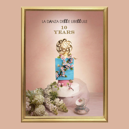 A framed image of the Nobile 1942 LA DANZA DELLE LIBELLULE EDP features a two-tiered cake with florals and a gold sphere topper, celebrating 10 years amid flowers, a tea set, and soft pink gourmand-inspired decor.