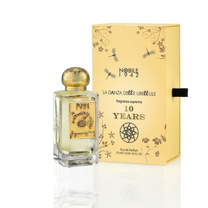 A clear glass bottle of LA DANZA DELLE LIBELLULE EDP - Nobile 1942 by Nobile 1942 with a gold label and ornate cap stands beside a yellow box. Both feature floral and dragonfly motifs, celebrating this gourmand fragrance's 10th anniversary.