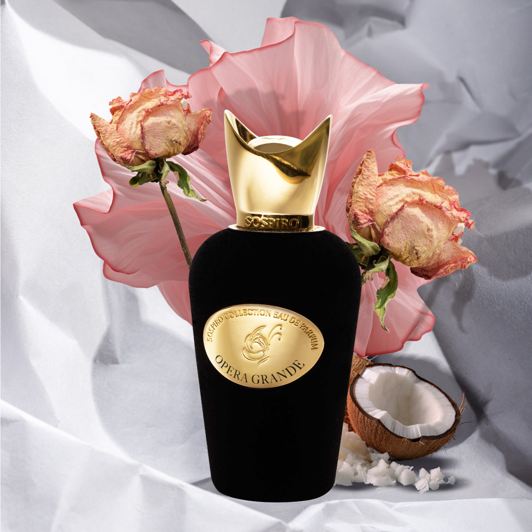 The Opera Grande Eau de Parfum by SOSPIRO is showcased in a black and gold bottle, set among pale pink flowers, dried roses, cracked coconut, scattered white petals, and accents of Turkish Rose.