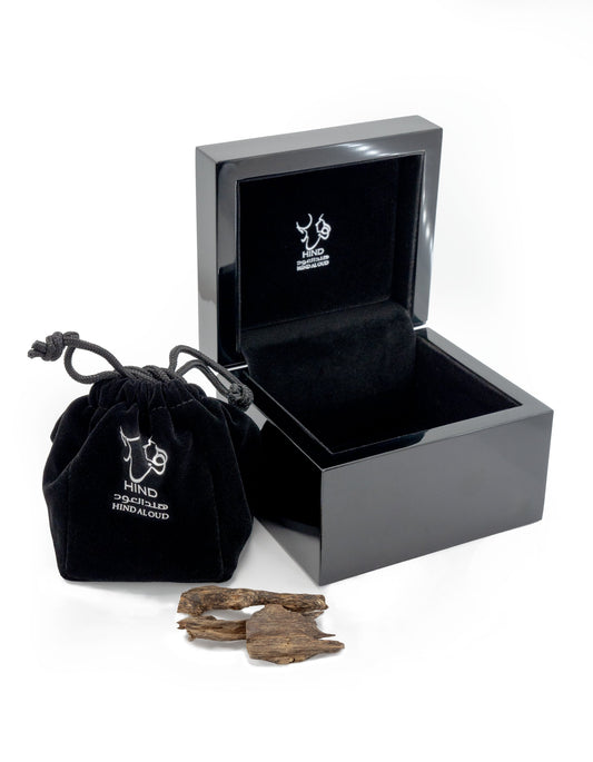 A glossy black jewelry box and a matching drawstring pouch, both with white Hind Al Oud logos, sit beside pieces of textured Oud Amiri Natural Oud, capturing the signature luxury of Hind Al Oud.