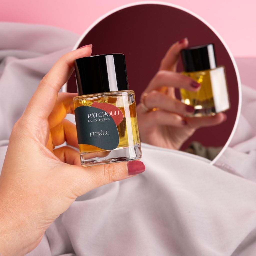A hand with red nail polish holds a bottle of Fennec Patchouli EDP, reflected in a round mirror against draped pink fabric, as its scent captures the mystique of a North African desert.