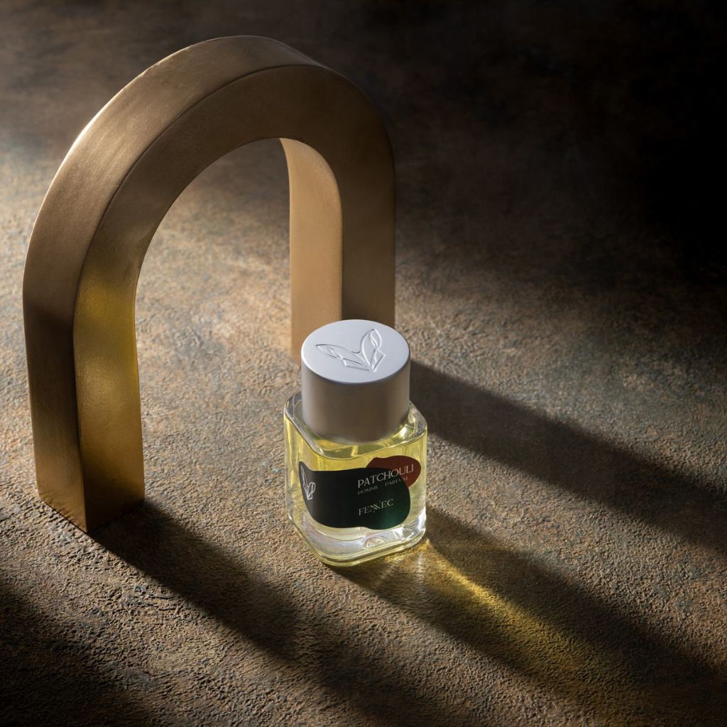 A glass Fennec Patchouli EDP bottle stands upright on a textured surface under soft light, with a metallic arch casting shadows in the background, evoking the mystique of patchouli across a North African desert.