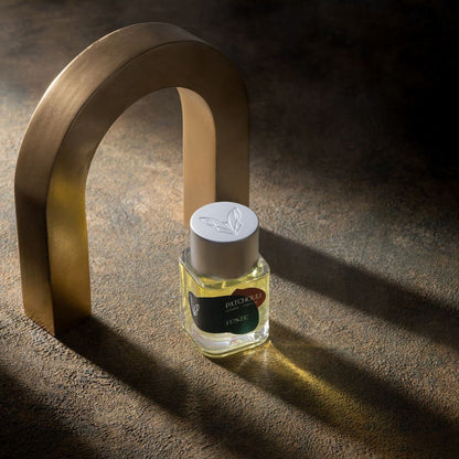 A glass Fennec Patchouli EDP bottle stands upright on a textured surface under soft light, with a metallic arch casting shadows in the background, evoking the mystique of patchouli across a North African desert.