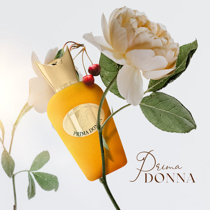 A yellow Prima Donna Eau de Parfum - Sospiro bottle by SOSPIRO, topped with a gold cap, sits among white roses, green leaves, and two red cherries. “Prima Donna” appears in elegant script, evoking the fragrance’s floral essence.