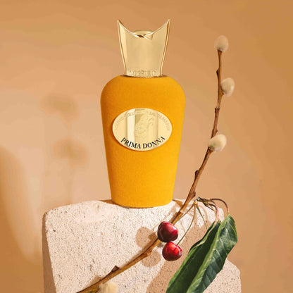 A yellow velvet Prima Donna Eau de Parfum by SOSPIRO, topped with a gold cap, rests on a stone block beside green leaves, red berries, and soft white buds—an elegant scene reflecting the floral essence of this fragrance.