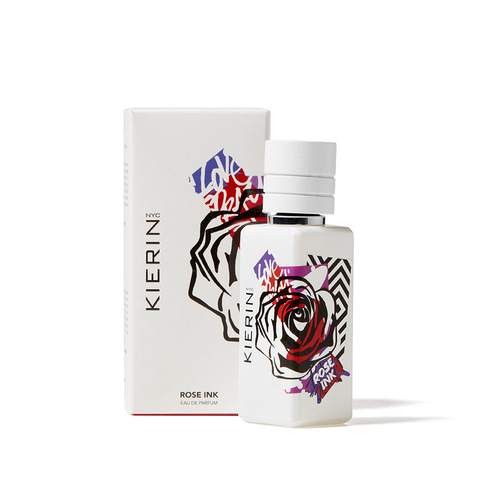 The Kierin Rose Ink Eau de Parfum, a unisex fragrance, comes in a white bottle and box with bold rose graphics and graffiti-inspired purple and red accents, highlighting its woodsy floral notes. Set shown on a white background.