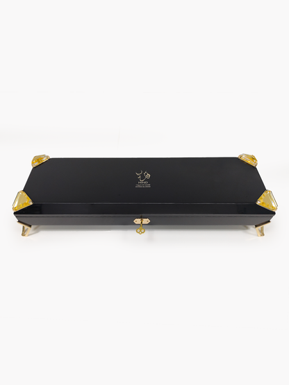 A sleek black rectangular box with gold accents, gold clasp, and gold feet features the Hind Al Oud logo and text on the lid, revealing it as the exclusive Royal Precious Collection perfume gift set by Hind Al Oud against a plain white background.