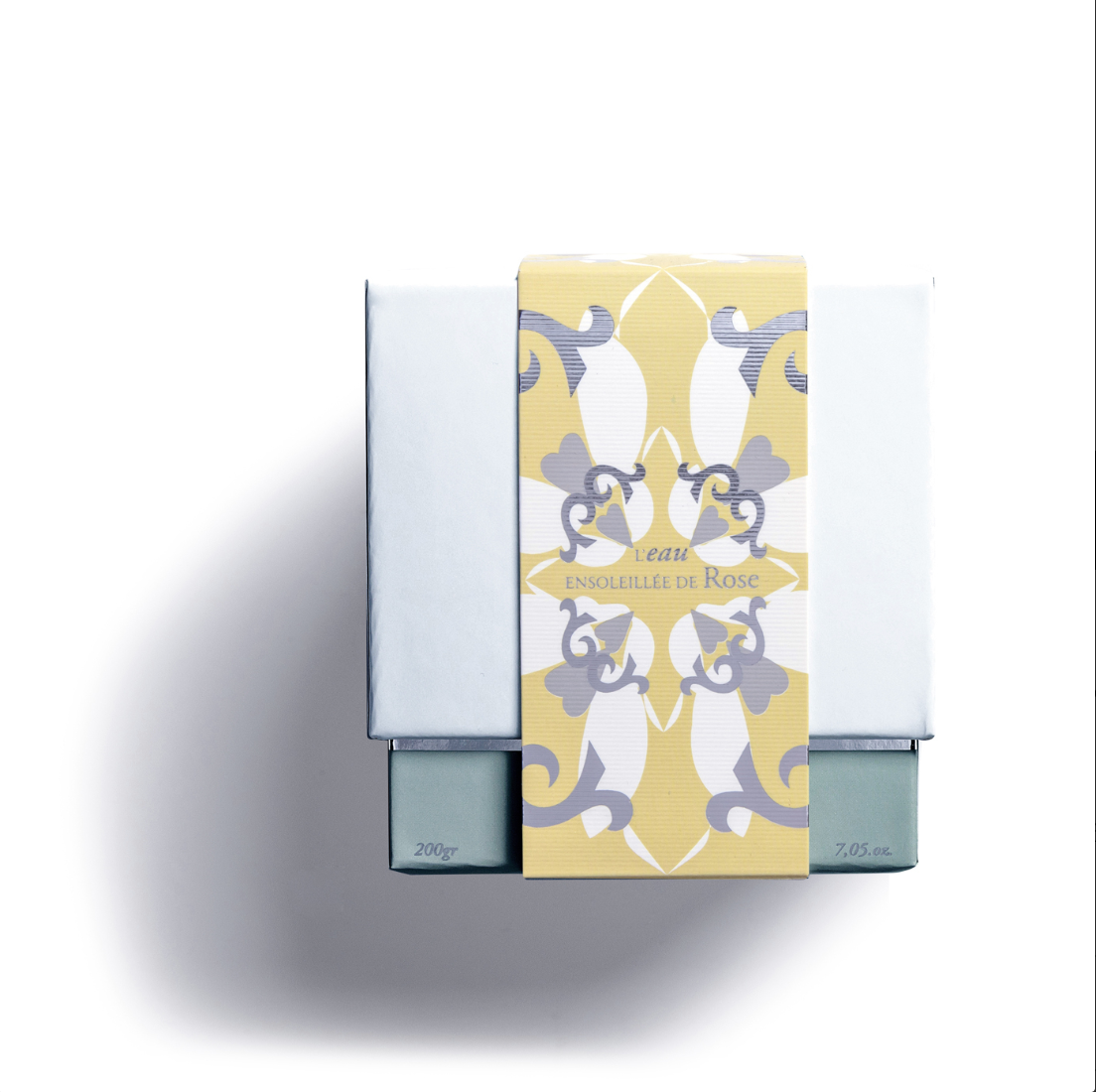 A white box with a yellow and gray floral label reading "L'EAU ENSOLEILLE DE ROSE," casts a shadow on a white background. It holds the ROSE'S SUN WATER SCENTED CANDLE by Rose Et Marius.