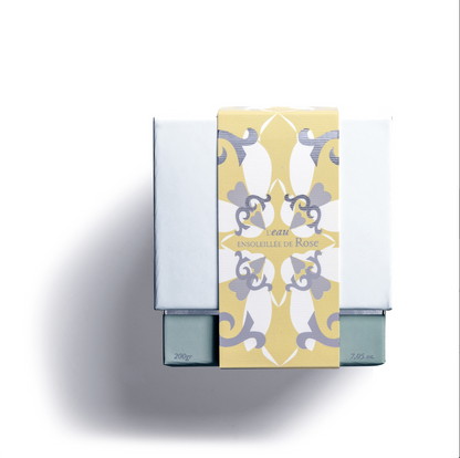 A white box with a yellow and gray floral label reading "L'EAU ENSOLEILLE DE ROSE," casts a shadow on a white background. It holds the ROSE'S SUN WATER SCENTED CANDLE by Rose Et Marius.