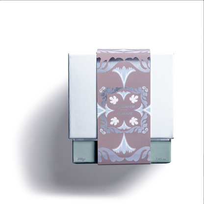 A white box with mauve, blue, and white floral bands and elegant text hints at the luxury Rose Et Marius AMBLING BENEATH THE ORATORY "SCENTED CANDLE UNE FLÂNERIE SOUS L'ORATOIRE", casting a soft shadow on a white background.