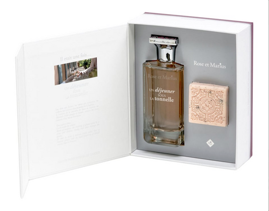 An open box reveals the Rose Et Marius GIFT SET ROSÉ WINE UNDER THE ARBOUR "UN VIN ROSÉ SOUS LA TONNELLE," featuring a unisex perfume bottle and ornate pink soap, with text on the inside cover.
