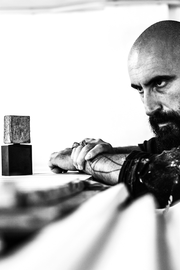 A contemplative black-and-white photo shows a bearded, bald man leaning on a table, gazing at a stone block atop a black cube—mirroring the earthy calm evoked by Filippo Sorcinelli’s UNUM OPUS 1144 "piece 1144" Eau de Parfum.