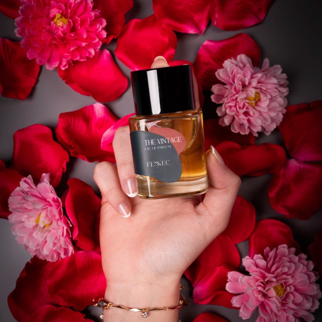 A hand holds a perfume bottle labeled The Vintage EDP by Fennec over a dark surface with pink and red petals, capturing the mysterious allure of North Africa and fragrance notes inspired by the elusive fennec fox.