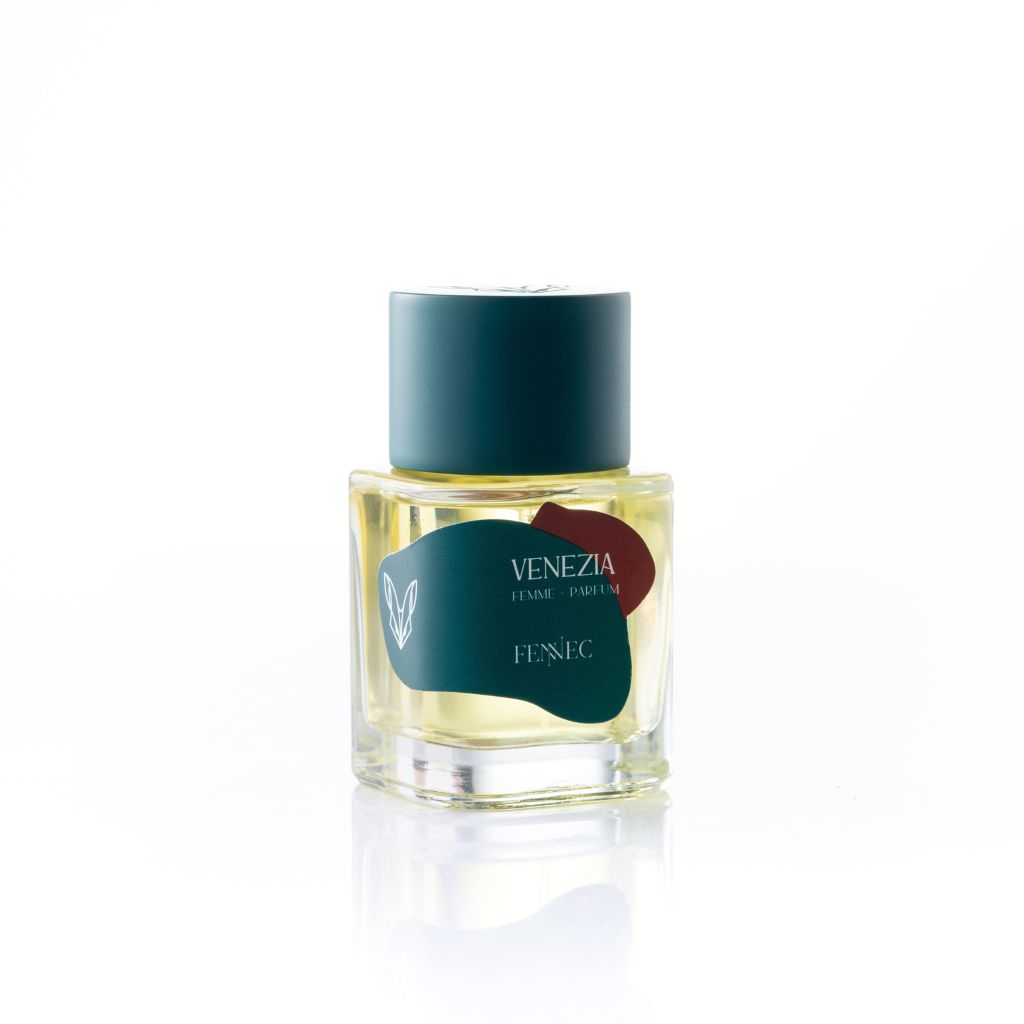 A clear glass bottle of Fennec Venezia Parfum features yellow liquid, a teal cap, and a teal label with red accents—all set on a white background. Experience perfume notes as alluring as the fennec fox.
