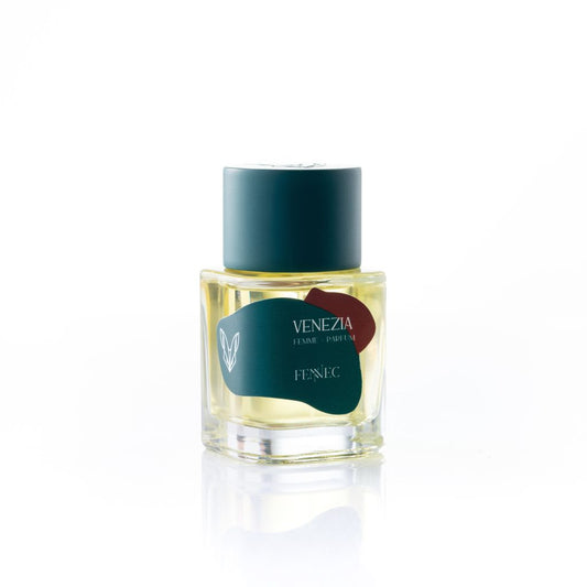 A clear glass bottle of Fennec Venezia Parfum features yellow liquid, a teal cap, and a teal label with red accents—all set on a white background. Experience perfume notes as alluring as the fennec fox.