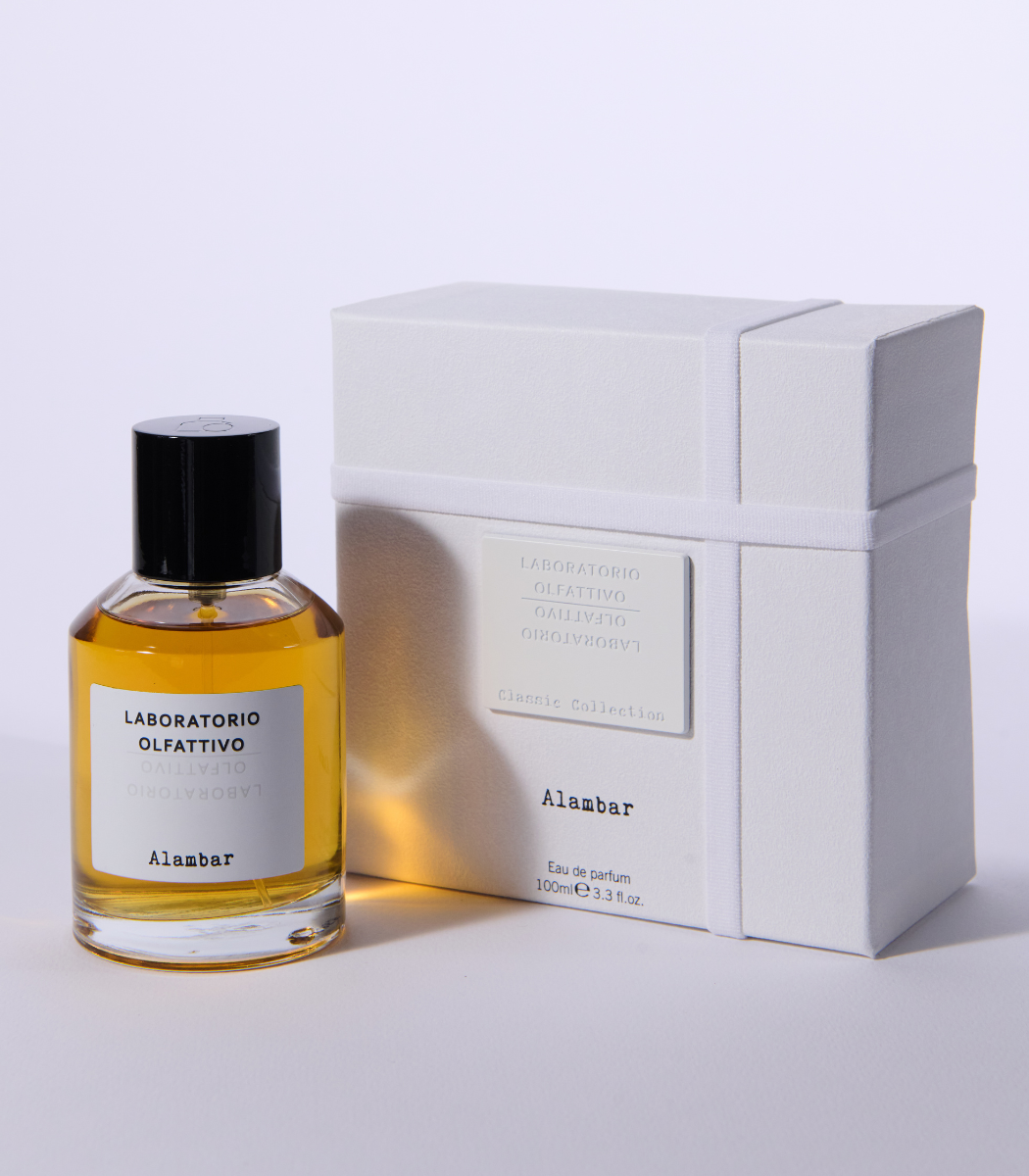 A bottle of Laboratorio Olfattivo Alambar perfume with a black cap sits beside its white textured box on a white surface, showcasing its amber-hued fragrance from Laboratorio Olfattivo.