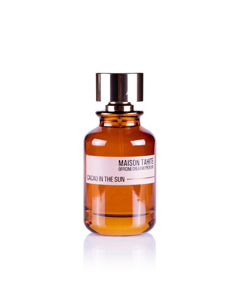 A brown glass bottle with a metallic cap, labeled "Cacao in the Sun Eau de Parfum" by Maison Tahite, sits on a white background, evoking a rich gourmand scent with distinctive cocoa notes.
