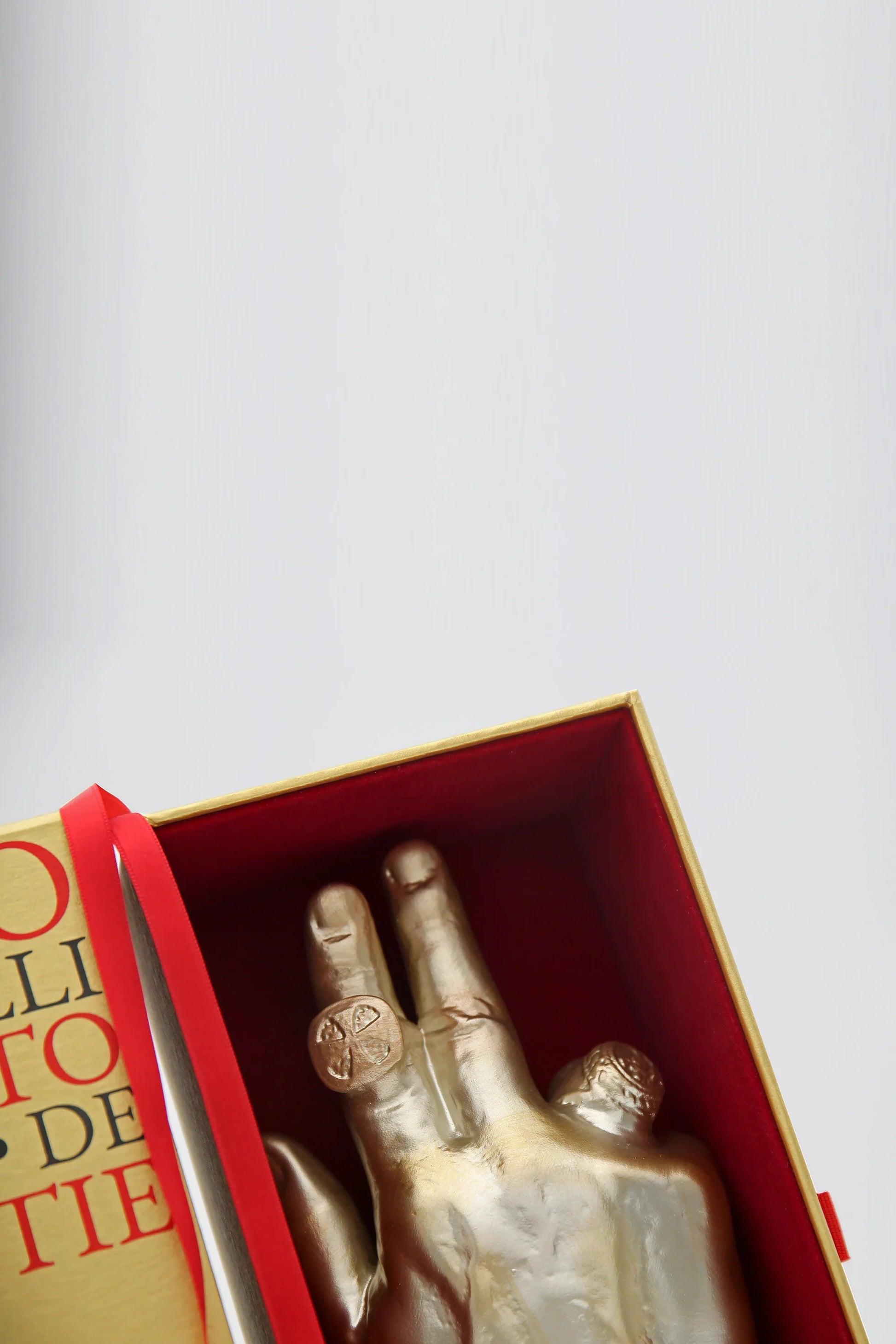 A gold-toned hand sculpture with rings sits inside a red-and-gold box beside a book’s edge and a silver knife, reflecting warm amber hues. PONT. MAX. Eau de Parfum by Filippo Sorcinelli is artfully displayed against a light, plain background.