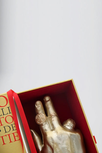 A gold-toned hand sculpture with rings sits inside a red-and-gold box beside a book’s edge and a silver knife, reflecting warm amber hues. PONT. MAX. Eau de Parfum by Filippo Sorcinelli is artfully displayed against a light, plain background.