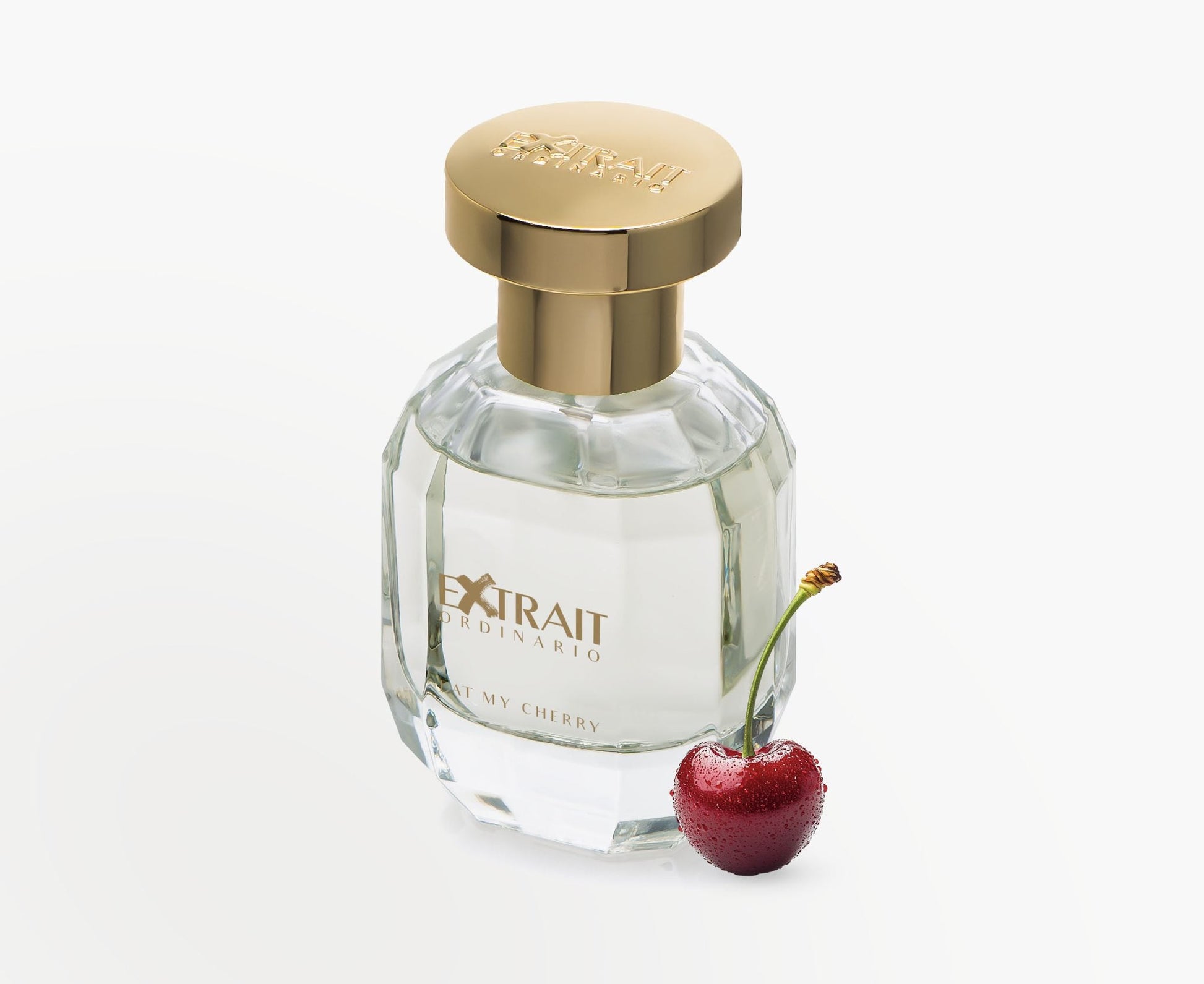 A clear glass bottle of Eat My Cherry - Extrait Ordinario by Extrait Ordinario, with a gold cap, stands beside a frosted cherry—a tempting invitation to experience this sensual fragrance against a crisp white background.