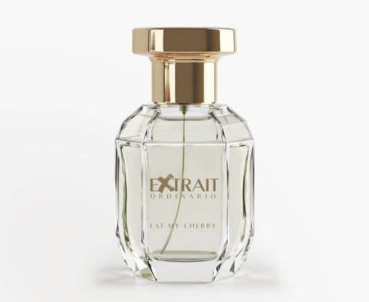 A clear, faceted glass bottle holds Extrait Ordinario’s Eat My Cherry perfume, featuring a gold cap and gold-lettered label. The pale cherry fragrance appears against a white background.