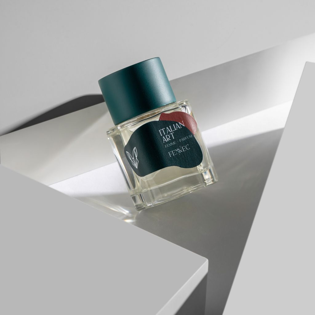 A glass bottle of Fennec's Italian Art Parfum with a green cap and label sits on a geometric white surface, its design inspired by the North African fennec fox, highlighted by soft light and sharp angles.