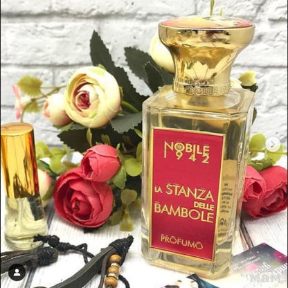 A bottle of LA STANZA DELLE BAMBOLE Eau de Parfum by Nobile 1942 stands before artificial red and pink roses, evoking the charm of a doll room. Nearby, other perfumes and items rest on a white surface reminiscent of playful childhood scenes.