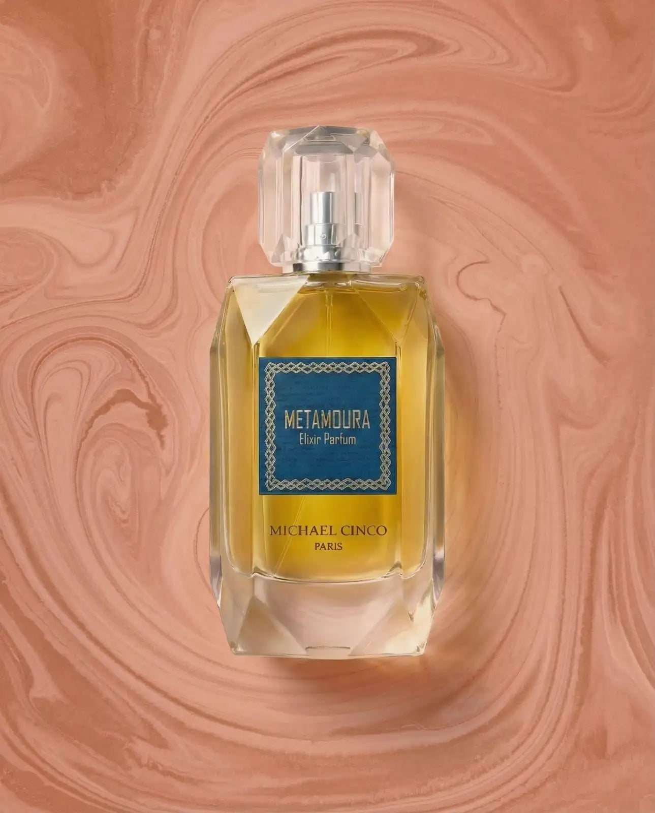 Michael Cinco Metamoura Blue Parfum in a faceted clear glass bottle with a blue label, set against a peach-colored marbled background.