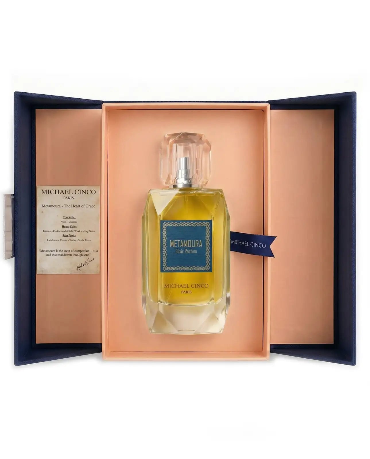 Michael Cinco Metamoura Eau de Toilette bottle presented inside an open peach-colored gift box with a navy blue outer flange. The clear glass bottle contains a golden-yellow liquid and features a blue label with white text.
