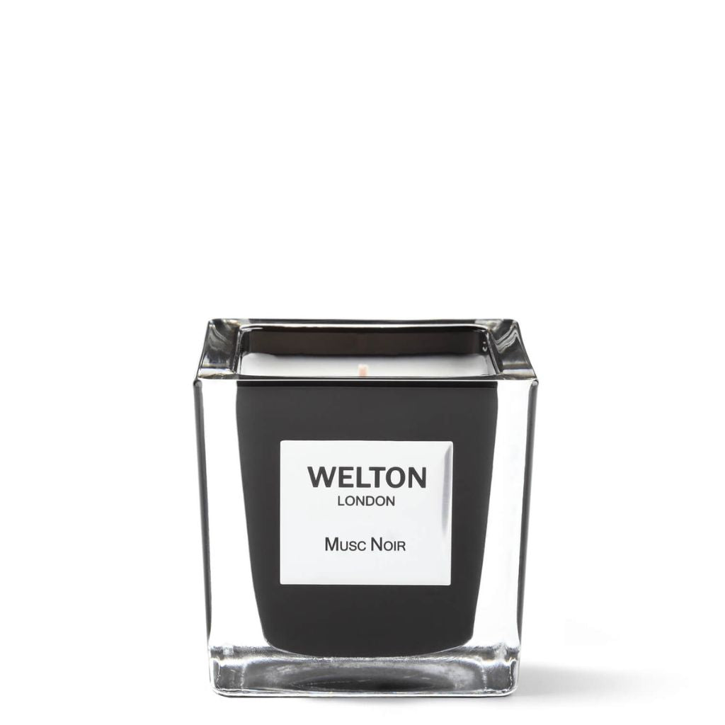 A square glass candle holder with a black candle inside, labeled "Welton London MUSC NOIR." The MUSC NOIR SCENTED CANDLE Classic features an original luxury fragrance by Welton London.