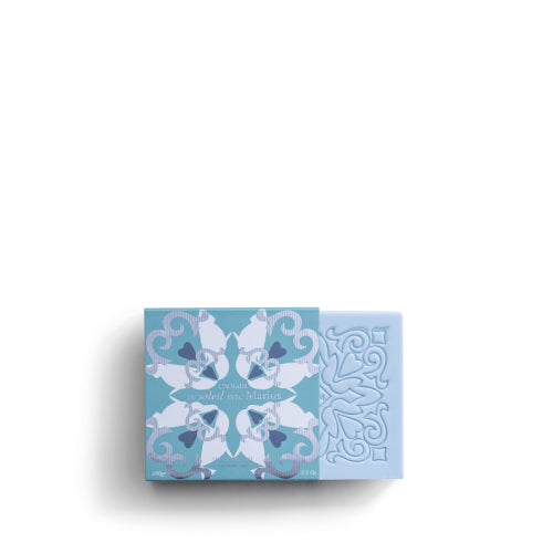 The ROSE ET MARIUS SCENTED SOAP by Rose Et Marius is presented in a light blue decorative box with floral patterns, revealing an intricate lid design. The French-made soap contains grape seeds for exfoliation and is perfumed naturally.