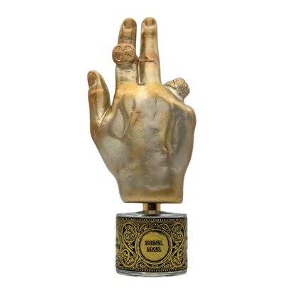 A golden hand sculpture with two raised fingers and a ring sits on an ornate black and gold base labeled BONNE MAIN, evoking amber and resin incense. Inspired by PONT. MAX. Eau de Parfum from Filippo Sorcinelli.