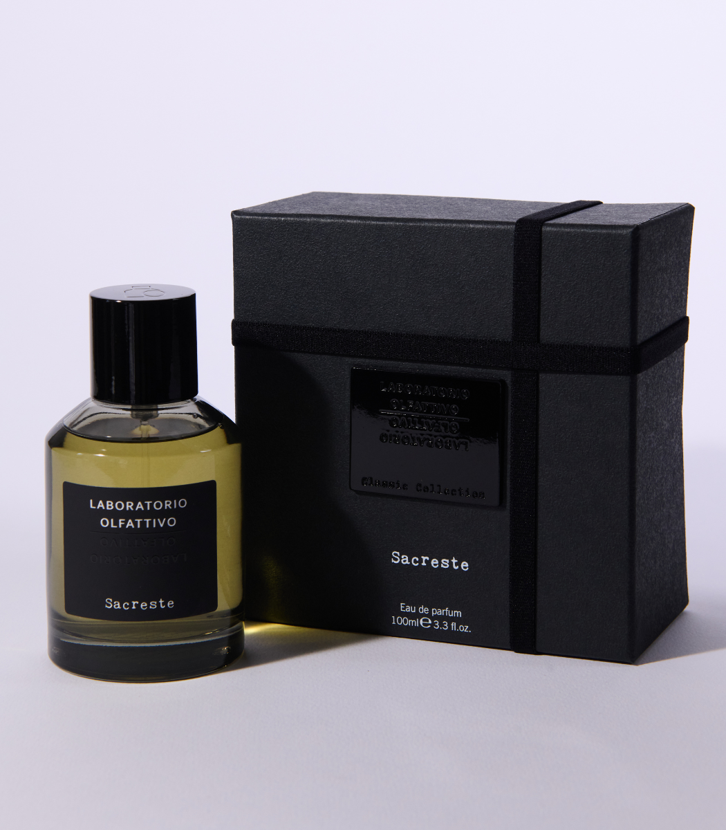 A bottle of Sacreste - Laboratorio Olfattivo stands beside its black rectangular box, both with minimalist black labels featuring white and silver text. The box is wrapped in a matching black ribbon.