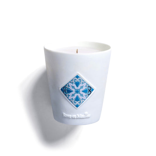 The BASKING IN THE SUN WITH MARIUS SCENTED CANDLE “UN BAIN DE SOLEIL AVEC MARIUS” by Rose Et Marius is a handmade French candle in a smooth white holder, featuring a blue and white tile design on the front and shown unlit against a plain background.