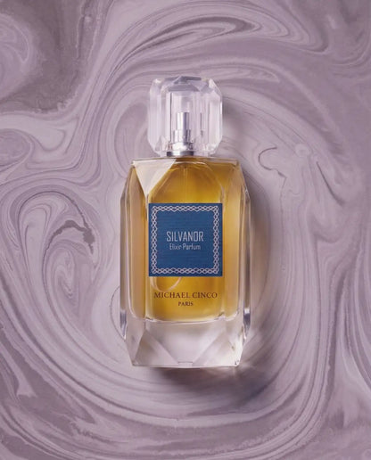 A bottle of Michael Cinco Silvandor Eau Parfum with golden-yellow liquid and a blue label, centered on a purple marbled background.