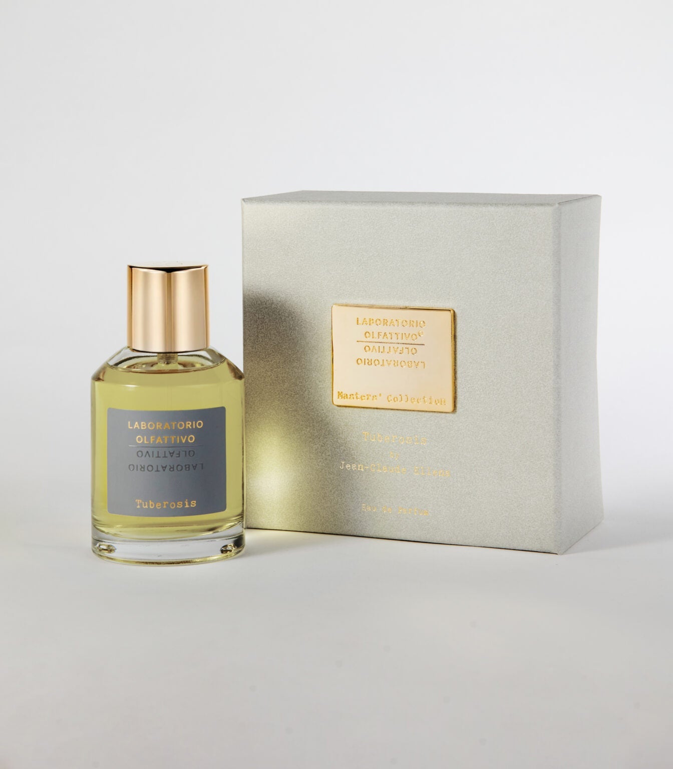 A clear glass bottle labeled Tuberosis - Laboratorio Olfattivo with a gold cap stands next to a silver, gold-accented box on a white background—its scent recalls tuberose flowers and the charm of an autumn evening.