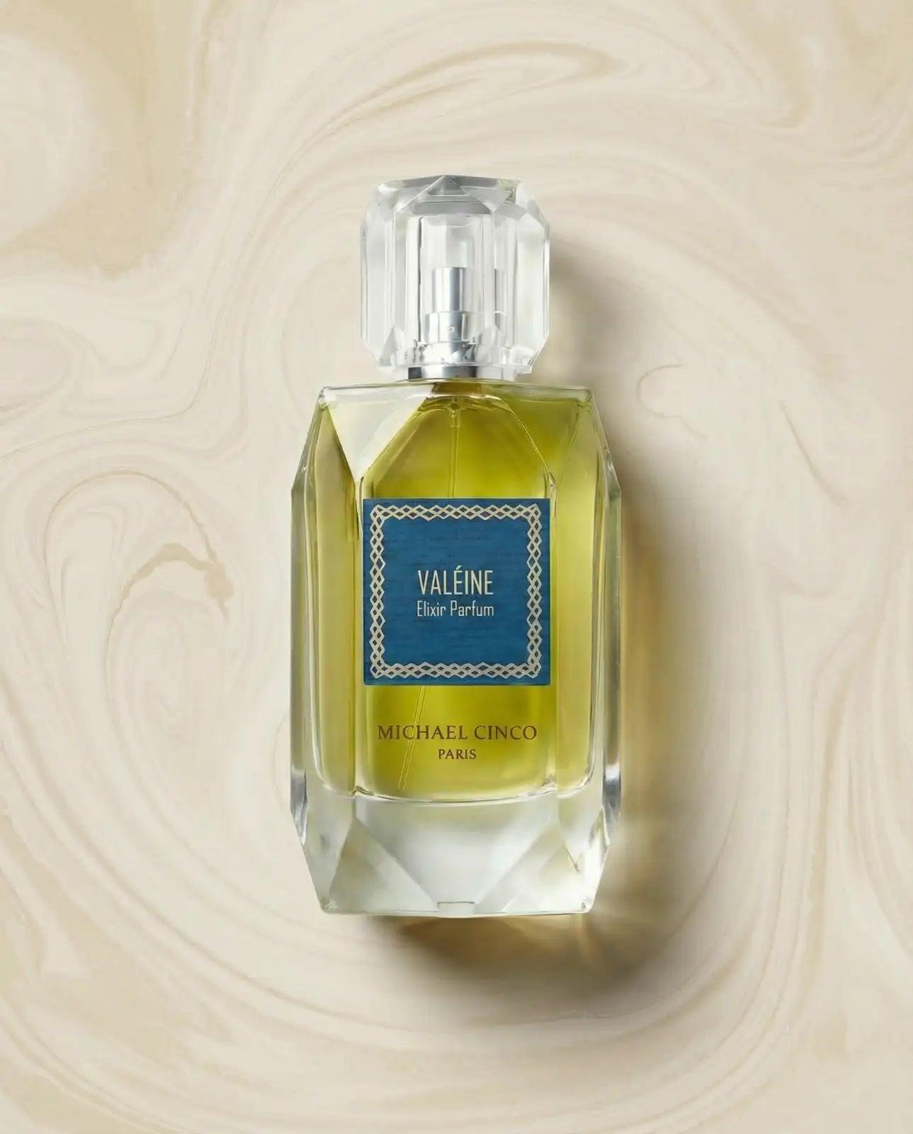 'Valéine' by Michael Cinco bottle, featuring a faceted clear glass design with a yellow-green liquid, a blue label, and a clear cap, set against a beige marbled background.