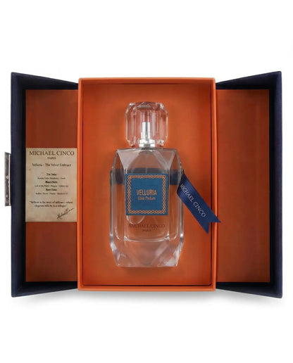 Michael Cinco Velluria Eau de Parfum in a clear glass bottle, presented inside an open orange and navy blue gift box with a brand card on the left.