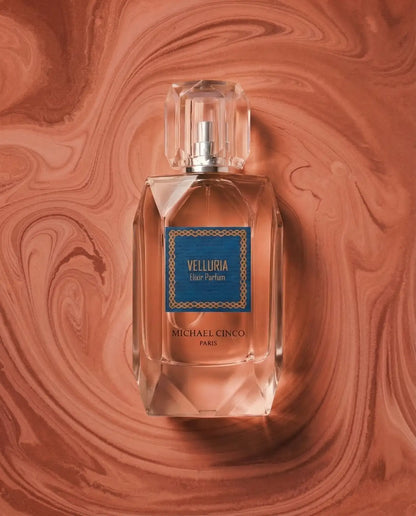 'VELLURIA Eau Parfum MICHAEL CINCO PARIS'bottle centered on a swirling terracotta-colored liquid background. The clear glass bottle shows the pale pink fragrance liquid inside, with a blue and gold label on the front and a clear faceted cap.