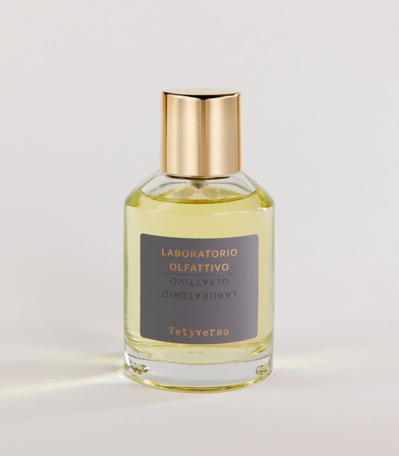 A clear glass bottle of Laboratorio Olfattivo Vetyverso TESTER perfume with a gold cap contains light yellow liquid. The label features gold and white text on gray. The bottle rests on a plain, light surface.